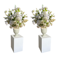 Two matching artificial flower arrangements in white urn-shaped vases, displayed on white rectangular pedestals against a plain white background. Saratoga Lake George New York Floral Rentals